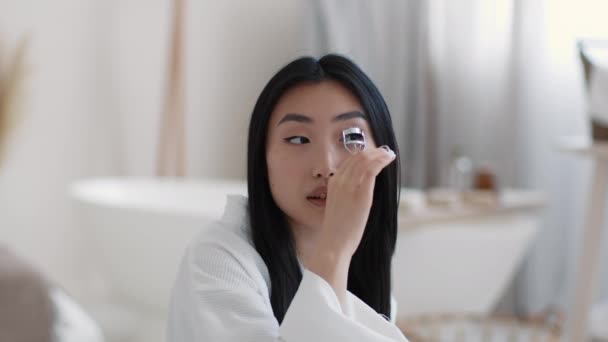 Young asian woman curling her eyelashes, using special curler, sitting at bathroom interior in morning, slow motion — Stockvideo