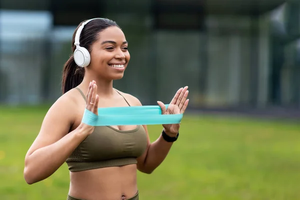 Adatto a giovani donne nere che si allenano con elastico, ascoltando musica in cuffie wireless durante l'allenamento all'aperto — Foto Stock