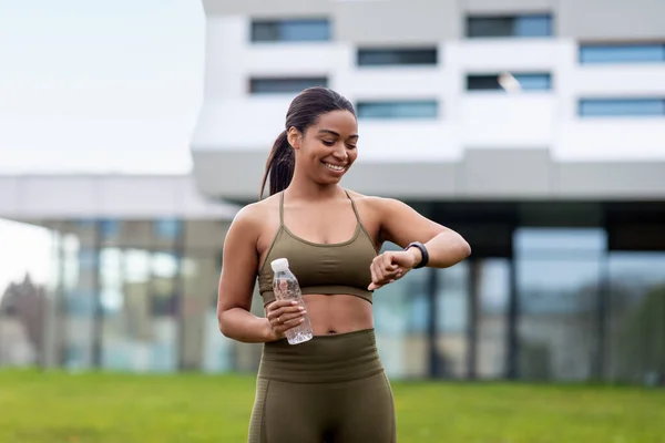 Retrato de esportivo jovem mulher negra com garrafa de água verificando seu smartwatch ou rastreador de fitness ao ar livre — Fotografia de Stock