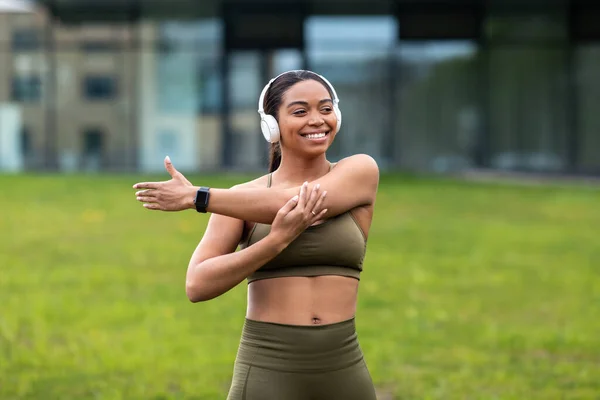 Giovane donna nera allegra che ascolta musica in cuffia, allunga il braccio, fa sport nel parco urbano — Foto Stock