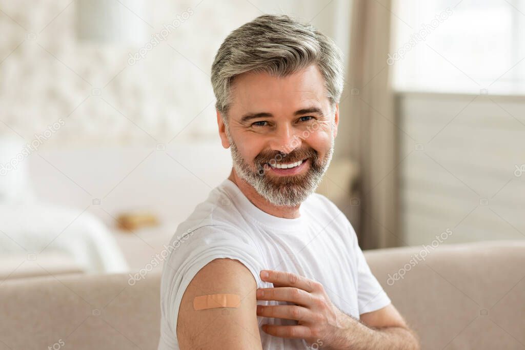 Joyful Vaccinated Man Showing Arm With Adhesive Bandage Sitting Indoors