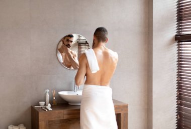 Kendine güvenen genç bir Arap aynaya bakıyor, sakala dokunuyor.