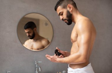 Beyaz havluya sarılmış bir adam elinde kozmetik ürünler püskürtüyor.