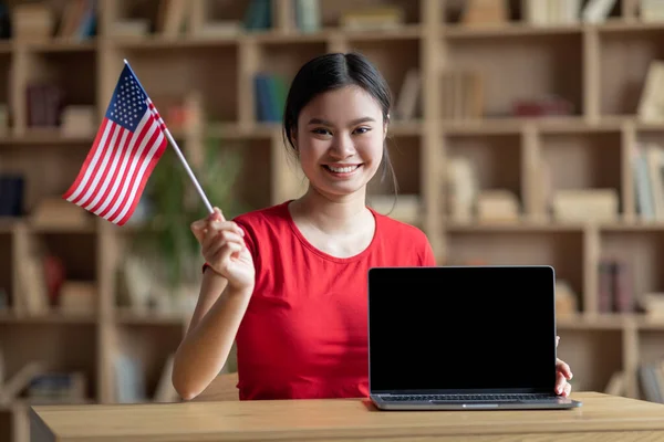 Glada unga japanska kvinnliga student hålla flagga USA, studerar med bärbar dator med tom skärm i vardagsrummet — Stockfoto
