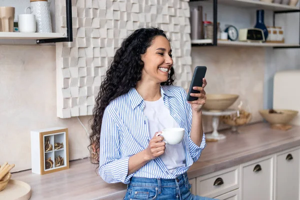 Mujer latina emocionada usando teléfono inteligente mientras bebe té de la mañana en la cocina en casa, leyendo mensajes y sonriendo — Foto de Stock