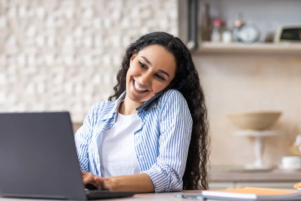 Conceito de negócio remoto. Retrato de senhora latina feliz falando no smartphone e digitando no laptop, sentado na cozinha — Fotografia de Stock