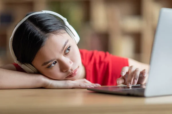 Smutný unavený mladý číňan dívka student v bezdrátových sluchátek leží na stole, dívá se na notebook — Stock fotografie
