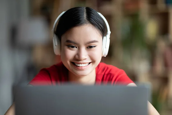 Triste joven estudiante chino cansado en auriculares inalámbricos se encuentra en la mesa, mira a la computadora portátil en la habitación —  Fotos de Stock
