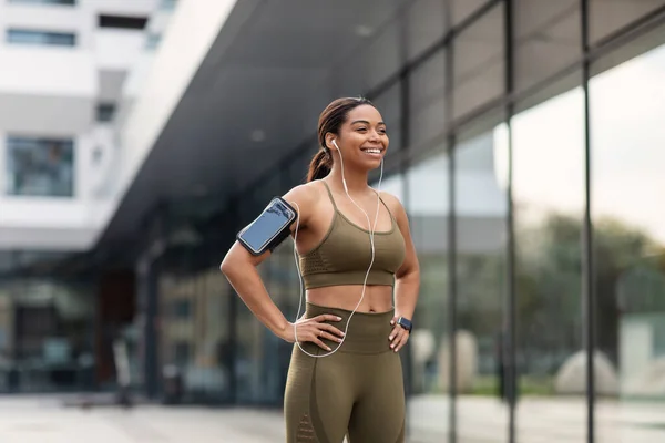 Giovane donna nera in abito sportivo ascoltare musica in cuffia, utilizzando il dispositivo mobile durante il jogging sulla strada della città — Foto Stock