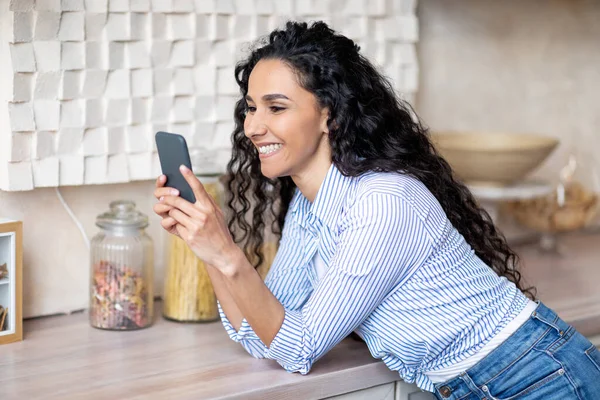 Mujer latina feliz en el uso casual de teléfonos inteligentes, navegar en las redes sociales, charlar con amigos, pasar tiempo en la cocina — Foto de Stock