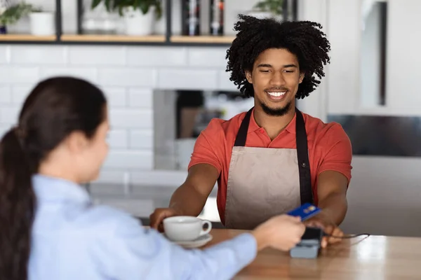 Lächelnd lockiger junger schwarzer Mann Barkeeper in Schürze am Bartresen gibt Terminal an Kundin mit Kreditkarte — Stockfoto
