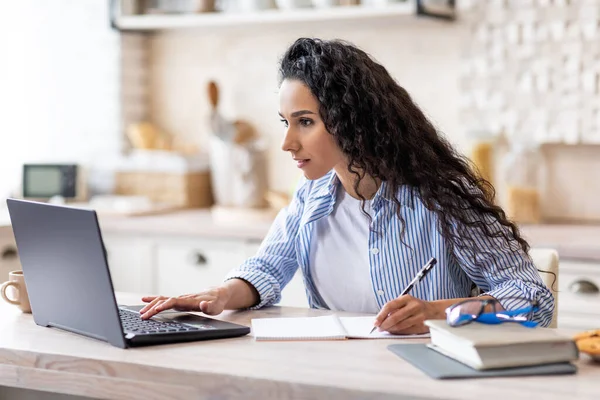 Koncentrált nő alkalmi dolgozik laptop minimalista konyha belső, nézi a képernyőn, és jegyzetel — Stock Fotó