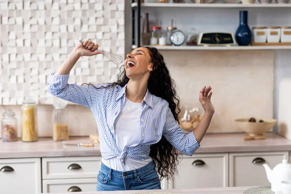 Overjoyed young lady singing at appliance as imaginary microphone, dancing in kitchen interior — Stock Photo, Image