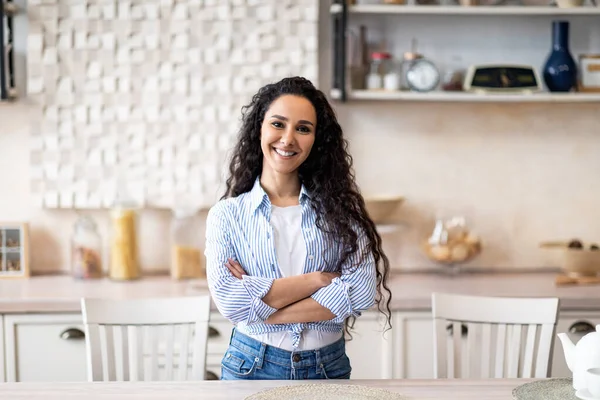 Portret van vrolijke latijnse huisvrouw poserend met gevouwen armen in de keuken, lachend en kijkend naar de camera, vrije ruimte — Stockfoto