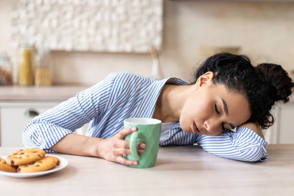Tired latin woman with coffee cup, waking up early and sleeping on table in light kitchen interior, free space