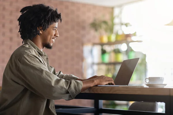 Feliz sonriente rizado joven negro mecanografía masculina en el ordenador portátil en la mesa en el interior de la cafetería moderna, sol destello, perfil —  Fotos de Stock