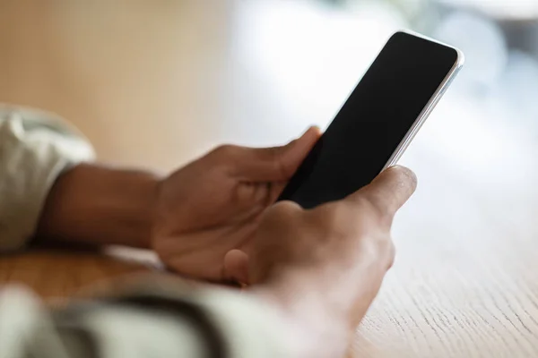 Millennial black male typing at smartphone with blank screen at wooden table in cafe interior, unrecognizable — Stock Photo, Image