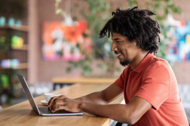 Gülümseyen kıvırcık, çekici siyah erkek bilgisayarda yazı yazıyor, kafenin içindeki sosyal ağlarda sohbet ediyor.