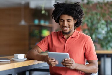 Mutlu, kıvırcık, yakışıklı, siyahi adam modern kafenin içinde tablete yazı yazıyor.