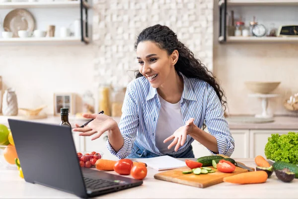 Счастливая латиноамериканка разговаривает с веб-камерой ноутбука во время приготовления свежего салата в кухонном интерьере, имея видеозвонок — стоковое фото
