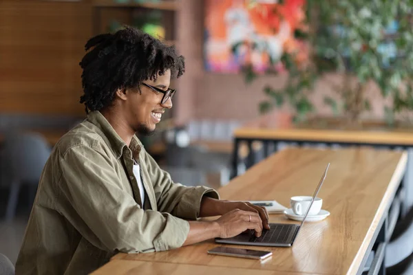 Fröhlich lächelnder Lockenkopf mit schwarzer Brille tippt auf Laptop am Tisch im Café, Kopierraum — Stockfoto