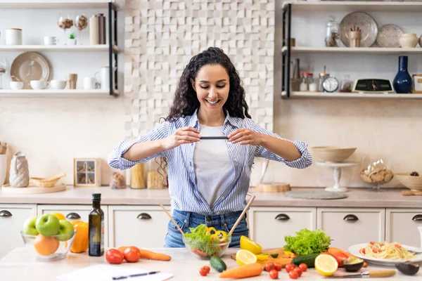 Красивая латинская блоггер делает фото салата для пищевого блога, стоя в кухонном интерьере, свободного места — стоковое фото