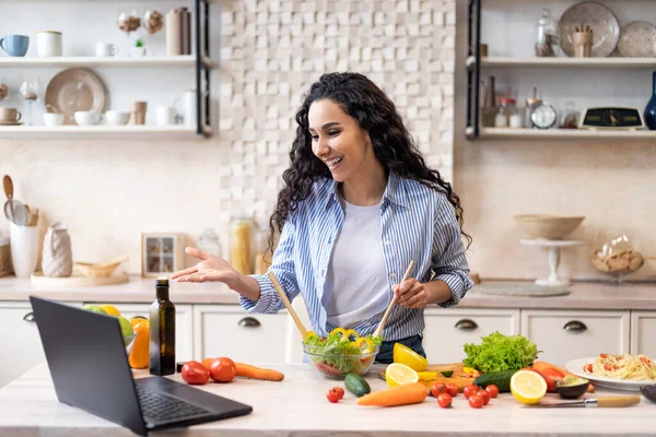 Online communication concept. Happy latin woman preparing salad and talking on video chat via laptop in kitchen — Stock Photo, Image