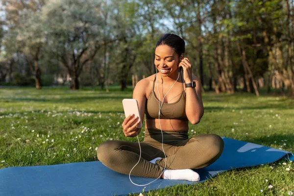 Tutta la lunghezza della giovane donna di colore allegra usando il cellulare, ascoltando musica durante l'allenamento di yoga all'aperto, spazio libero — Foto Stock