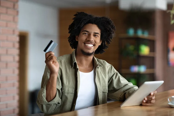 Alegre sonriente rizado milenario negro hombre celebrar tarjeta de crédito y tableta en la mesa de madera en línea de compras en la cafetería —  Fotos de Stock