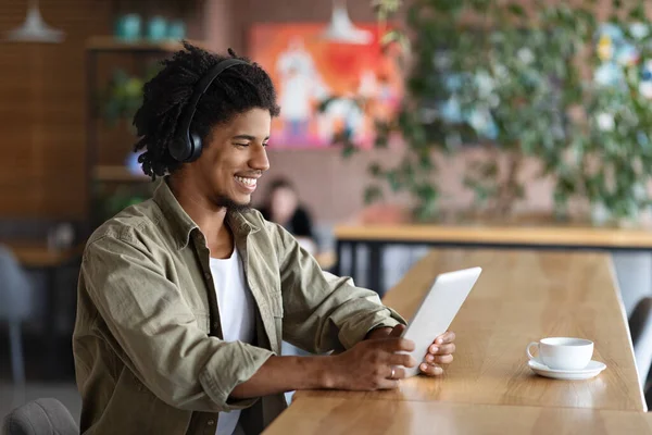 Feliz sonriente rizado joven hombre negro en auriculares inalámbricos mirar a la tableta y escuchar música en la cafetería —  Fotos de Stock