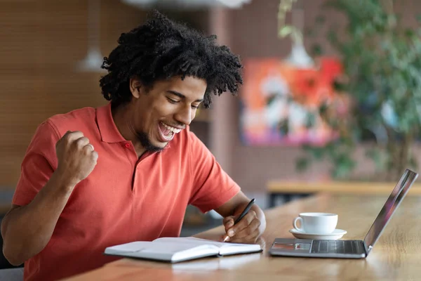 Felice felice eccitato riccio bello giovane uomo nero prendere appunti nel quaderno, gesto di vittoria in caffè interno — Foto Stock