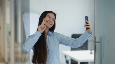 Sosyal medya içeriği. Genç oyuncu Asyalı bayan yönetici cep telefonuyla selfie çekiyor, ofisteki cihaza gülümsüyor.