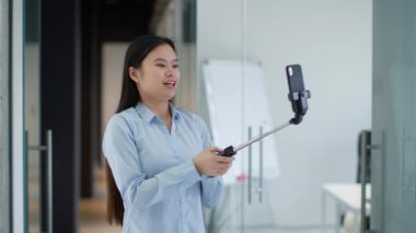 Genç Asyalı blog yazarı akıllı telefondan video kaydediyor, el sallıyor ve cihazla konuşuyor, Monopod 'da cep telefonu taşıyor.