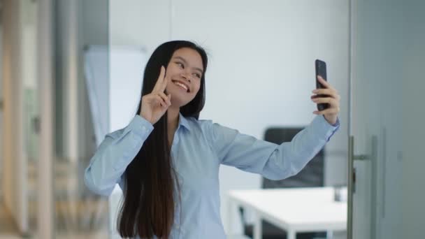 소셜 미디어 콘텐츠. 젊고 장난기많은 아시아 여자 매니저가 휴대 전화로 셀피를 만들고 사무실에 있는 기기에 미소를 짓고 있습니다 — 비디오