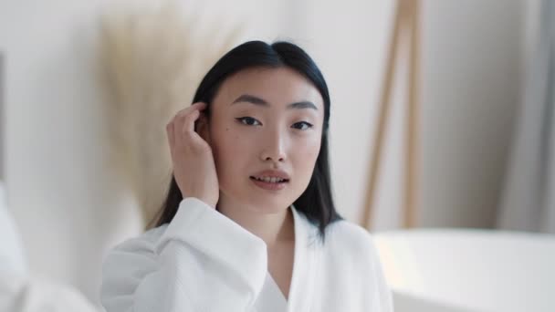 Retrato de belleza de una joven asiática bien arreglada arreglando el cabello y sonriendo a la cámara, sentada en el baño, cámara lenta — Vídeos de Stock