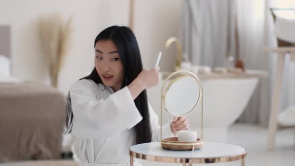 Young attractive asian lady sitting at bathroom in morning and combing her silky hair, looking at mirror, free space — Stock Video