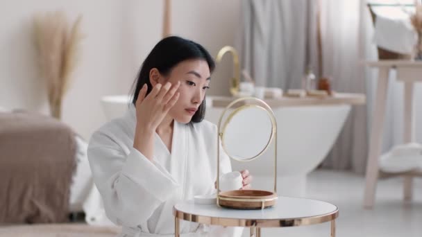 Rotina de beleza todos os dias. Jovem mulher asiática aplicando creme hidratante no rosto, olhando para o espelho, espaço livre — Vídeo de Stock