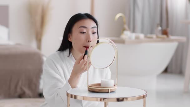 Mladý krásný asijské žena nanášení červené rtěnky na rty, dělat jasný make-up, sedí v rozmazané koupelně, volný prostor — Stock video