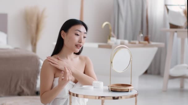 Young asian woman wrapped in towel applying pampering lotion on shoulders and neck, moisturizing body at bathroom — Stock Video