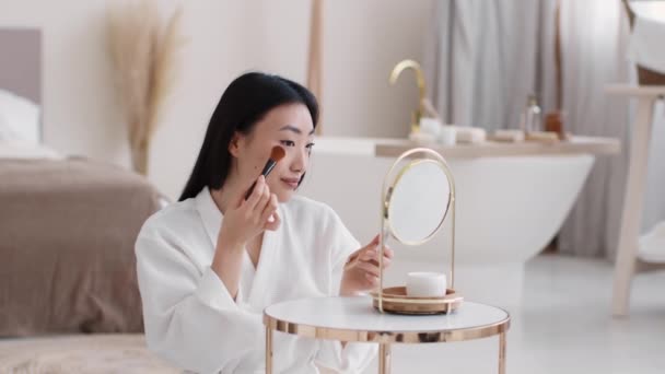Młoda ładna azjatycka kobieta robi sobie makijaż, nakłada róż na twarz, patrzy w lustro na wnętrze łazienki — Wideo stockowe