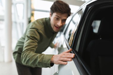 Mutlu genç adam hayallerinin yeni otomobiline dokunuyor, galeriden modern araba alıyor, seçici odaklanıyor.