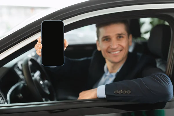 Glad ung bil säljare visar smartphone med tom skärm medan du sitter i ny bil på auto återförsäljare, mockup — Stockfoto