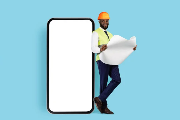 Black Smiling Construction Engineer With Blueprints Leaning At Big Blank Smartphone