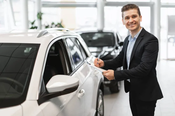 Happy young auto insurance agent standing near new car, checking it, taking notes, working at automobile dealership
