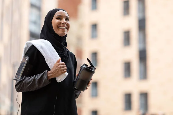 Wanita muslim muda yang senang mengenakan jilbab dan pakaian olahraga dengan telepon di bahu dengan cangkir koktail dan handuk di jalan — Stok Foto