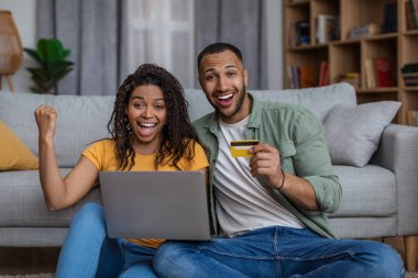Dizüstü bilgisayarı ve kredi kartıyla başarıyı kutlayan heyecanlı Afro-Amerikan çift online kazanca duygusal tepki veriyor.