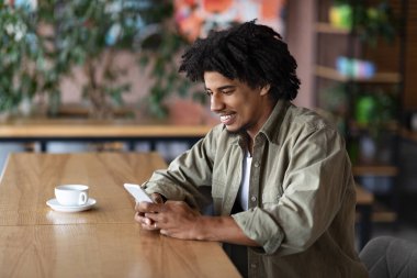 Mutlu kıvırcık saçlı, yakışıklı, bin yıllık siyahi adam modern kafenin içindeki masada akıllı telefona yazı yazıyor.