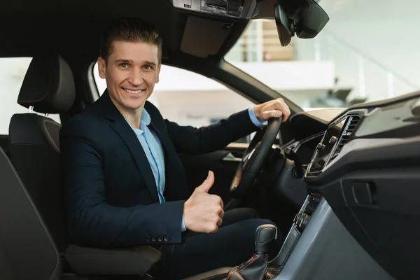 Szczęśliwy młody sprzedawca samochodów siedzi w nowym samochodzie, pokazując gest kciuka w górę, polecając nowoczesny salon — Zdjęcie stockowe