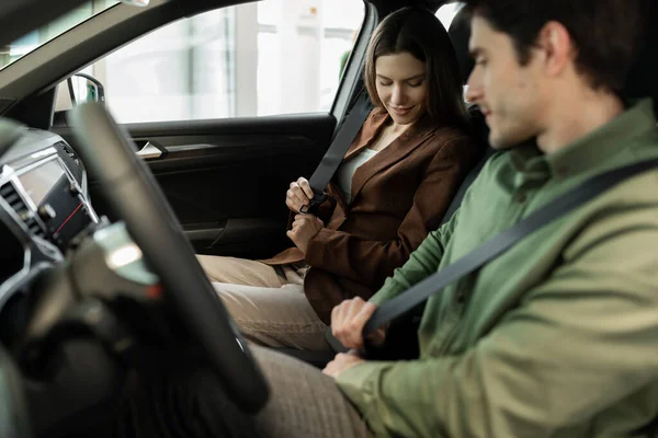 Positive junge Ehepartner überprüfen Neuwagen, sitzen im Auto, schnallen Sicherheitsgurte an, gehen auf Testfahrt im Autohaus — Stockfoto