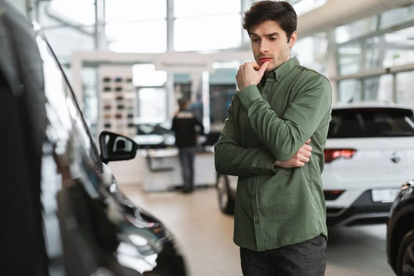 Umsichtiger junger Mann denkt nach, trifft die Wahl, hat Zweifel am Neuwagenkauf im Autohaus, kopiert Platz — Stockfoto